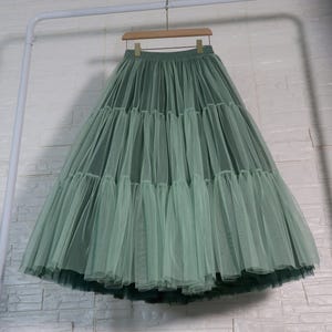 Peut inclure: Une jupe en tulle à étages vert sauge dotée d'une taille haute avec une ceinture élastique froncée. La jupe est composée de plusieurs couches de tissu en maille fine et transparente qui créent une silhouette volumineuse suspendue à un cintre.