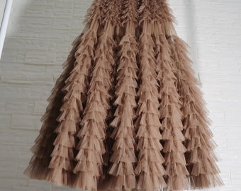 May include: A floor-length, brown tulle skirt with multiple layers of ruffles. The skirt is hanging on a white metal rack against a white brick wall. The skirt's elastic waistband is visible.