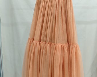 May include: A peach-colored tulle skirt hangs from a wooden hanger. The skirt has a gathered waistband and a tiered, flowing design. The fabric appears lightweight and semi-sheer, creating a soft, romantic look. The skirt is suitable for special occasions.