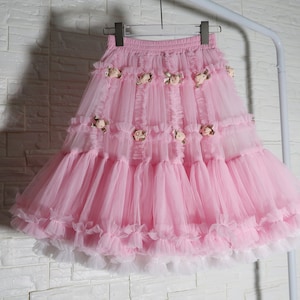 Puede incluir: Una falda de tul rosa con múltiples capas con volantes y pequeños detalles florales. La falda está colgada en un soporte blanco contra una pared de ladrillos blancos. La falda tiene un ribete blanco en la parte inferior.