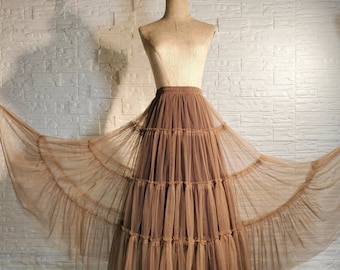 May include: A brown tulle skirt with multiple layers and a ruffled hem. The skirt is displayed on a mannequin against a white brick wall.