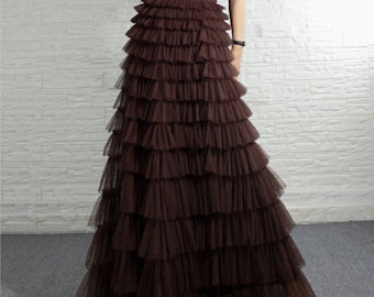 May include: A floor-length, tiered skirt in a rich brown hue. The skirt features multiple layers of ruffled fabric, creating a voluminous and flowing silhouette. The waistband is a darker shade of brown. The skirt is made of a sheer material.