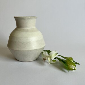 May include: A matte off-white ceramic vase with a wide, angular midsection and a flared rim. The vase features a subtle speckled texture throughout its surface. Beside the vase lies a single stem of white alstroemeria flowers with green leaves.