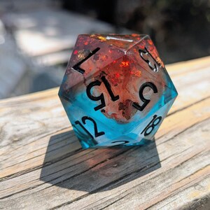 May include: A twenty-sided die featuring a dual-tone design with a translucent blue base and a reddish-orange upper section containing embedded glitter. The die has black engraved numbers, including 15, 5, 8, 12, and 7, clearly visible on its faces.