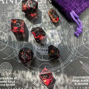 May include: A collection of polyhedral dice with a red and black marbled design, scattered on a surface with a skull graphic. A purple velvet dice bag is in the background. The dice are for role-playing games.