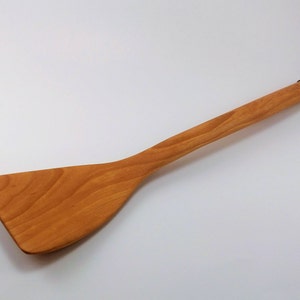 May include: A wooden spatula with a long handle and a wide, flat blade. The spatula is made of light brown wood and has a natural finish. It is a kitchen utensil used for flipping food.