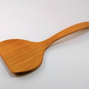 May include: A wooden spatula with a wide, rounded head and a long, thin handle. The spatula is made of light brown wood and has a smooth, polished finish.