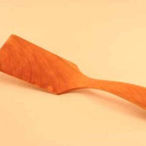 May include: A wooden spatula with a long, curved handle. The spatula is made of light brown wood with a natural grain pattern.