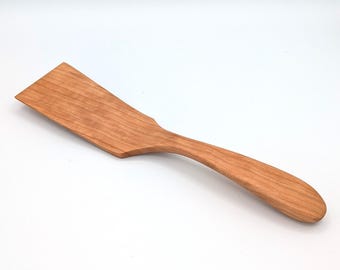 May include: A kitchen spatula crafted from light brown wood with a visible natural grain pattern. The utensil features a wide, flat, rectangular head that tapers into a smooth, rounded handle, designed for flipping and turning food items.