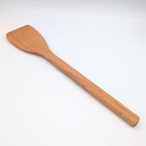 May include: A light brown wooden spatula with a long, cylindrical handle and a wide, flat, angled head. The kitchen utensil features a smooth, natural wood grain pattern throughout its surface, showcasing a consistent finish and simple design.