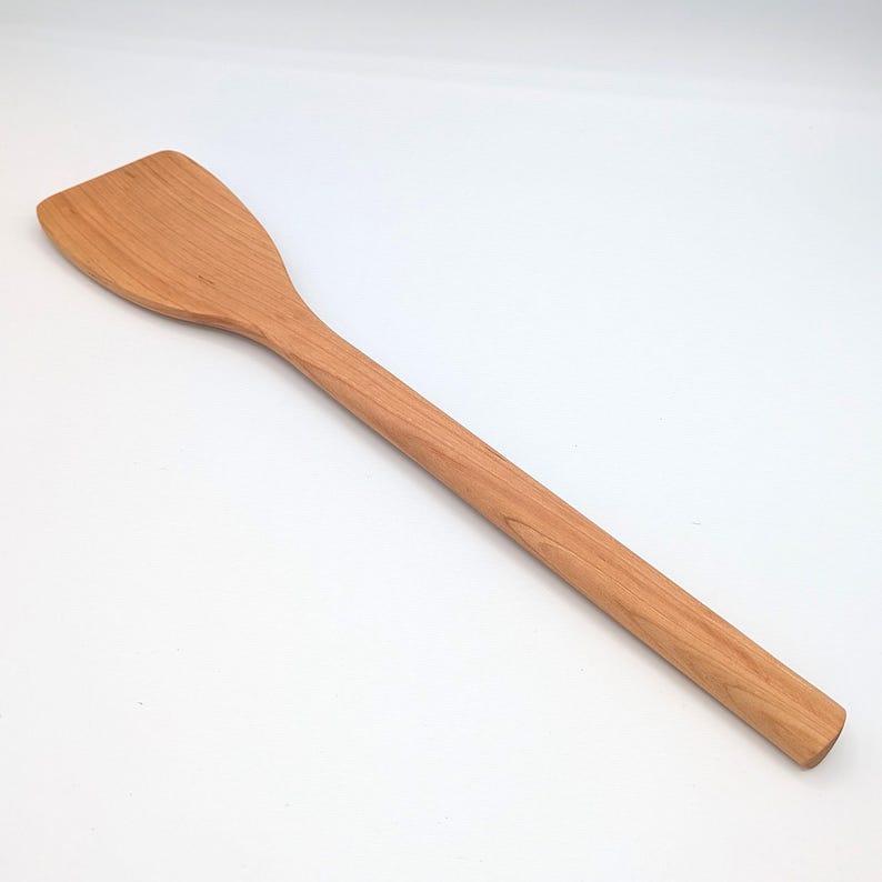 May include: A light brown wooden spatula with a long, cylindrical handle and a wide, flat, angled head. The kitchen utensil features a smooth, natural wood grain pattern throughout its surface, showcasing a consistent finish and simple design.