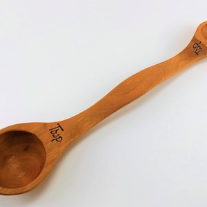 May include: A wooden spoon with two measuring cups, one marked "Tbsp" and the other marked "dtsp".