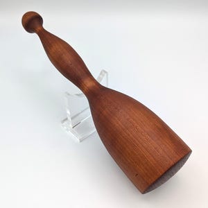 May include: A hand-turned wooden mallet with a smooth, reddish-brown finish and visible wood grain. The tool features a long, tapered handle ending in a rounded knob and a cylindrical head with a flat striking surface, crafted from solid wood.