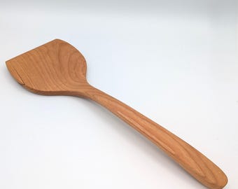 May include: A light brown wooden spatula with a wide, flat, angled head and a long, smooth, tapered handle. The natural wood grain is visible throughout the surface of the utensil, showing a consistent texture and a warm, medium-toned finish.