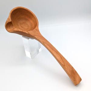 May include: A hand-carved wooden ladle with a smooth, light brown finish and visible wood grain patterns. The ladle features a deep, rounded bowl with a distinct side spout for pouring, connected to a long, curved handle that tapers toward the end.