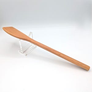 May include: A long, light brown wooden cooking spatula with a flat, angled head and a smooth, rounded handle. The grain of the wood is visible throughout the surface of the utensil, which is resting on a clear acrylic display stand.