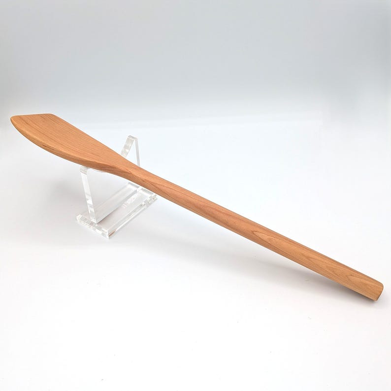 May include: A long, light brown wooden cooking spatula with a flat, angled head and a smooth, rounded handle. The grain of the wood is visible throughout the surface of the utensil, which is resting on a clear acrylic display stand.