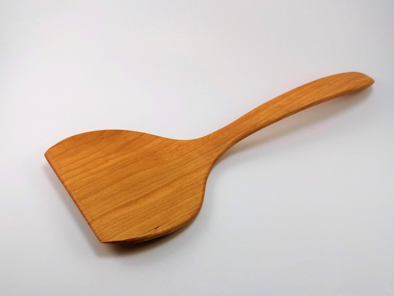 May include: A light brown wooden spatula with a wide, flat, and slightly curved head attached to a long, smooth, and tapered handle. The wood grain is visible across the entire surface, showing natural patterns and a warm, polished finish.