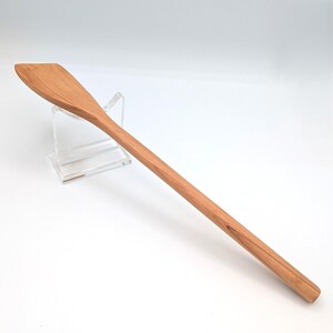 May include: A long, slender wooden spatula with a smooth, light brown finish and a distinct grain pattern. The utensil features a flat, angled head that tapers into a rounded, elongated handle, showcasing a natural wood texture throughout its length.
