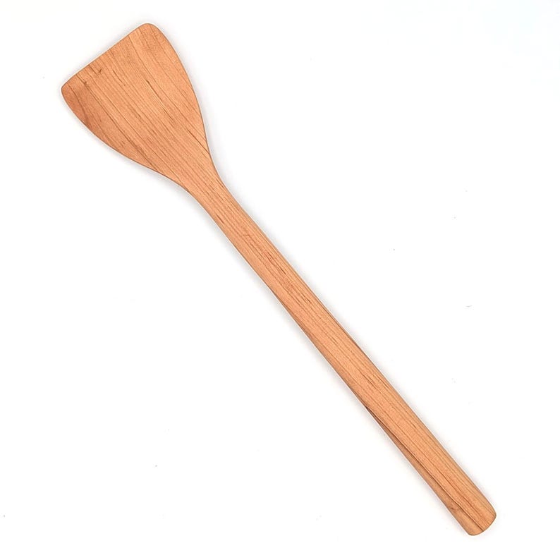 May include: A light brown wooden spatula with a long, straight handle and a wide, flat, angled head. The natural wood grain is visible throughout the surface of the utensil, showing a smooth texture and a consistent, warm, light brown color.