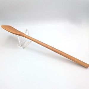 May include: A long, slender wooden cooking spoon with a light brown grain and a flat, angled head. The spoon features a smooth, rounded handle and is supported by a clear acrylic stand, showcasing the natural texture and elongated design.