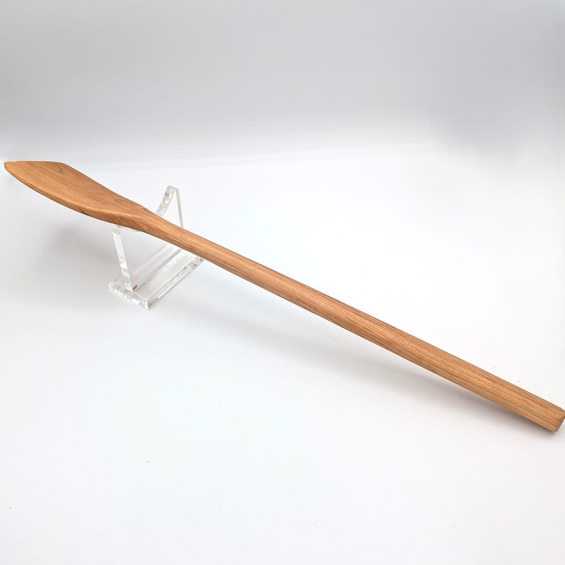 May include: A long, slender wooden cooking spoon with a light brown grain and a flat, angled head. The spoon features a smooth, rounded handle and is supported by a clear acrylic stand, showcasing the natural texture and elongated design.