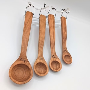 May include: A set of four wooden measuring spoons with a natural grain finish, each featuring a long handle and a round bowl. The spoons are labeled with black text as Tbsp, tsp, 1/2 tsp, and 1/4 tsp, and are suspended by black cords.
