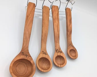 May include: A set of four wooden measuring spoons with a natural grain finish, each featuring a long handle and a round bowl. The spoons are labeled with black text as Tbsp, tsp, 1/2 tsp, and 1/4 tsp, and are suspended by black cords.