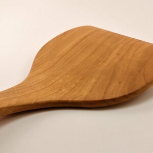 May include: A light brown wooden spatula with a wide, flat, and curved head. The kitchen utensil features a smooth, polished surface with visible natural wood grain patterns running along the length of the handle and the broad, rounded blade.