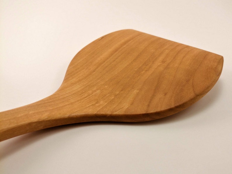 May include: A light brown wooden spatula with a wide, flat, and curved head. The kitchen utensil features a smooth, polished surface with visible natural wood grain patterns running along the length of the handle and the broad, rounded blade.