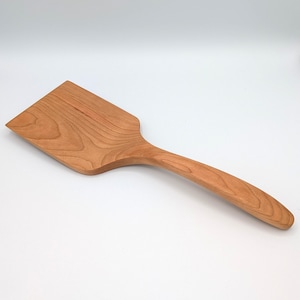 May include: A light brown wooden spatula with a wide, flat head and a smooth, curved handle. The kitchen utensil features a visible natural wood grain pattern throughout its surface, showcasing a warm, reddish-brown tone and a solid construction.