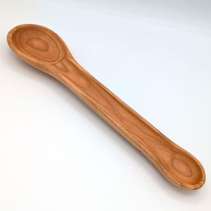 May include: A double-ended wooden spoon featuring a large, deep oval bowl on one end and a smaller, shallower circular bowl on the opposite end. The utensil displays a natural light brown wood grain pattern with a smooth, polished finish throughout.