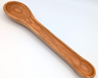 May include: A double-ended wooden spoon featuring a large, deep oval bowl on one end and a smaller, shallower circular bowl on the opposite end. The utensil displays a natural light brown wood grain pattern with a smooth, polished finish throughout.