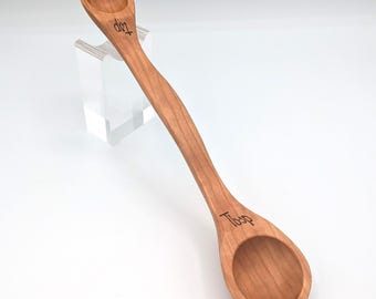 May include: A double-ended wooden measuring spoon with a light brown wood grain finish. The smaller bowl is engraved with the text tsp, and the larger bowl is engraved with the text Tbsp. The handle has a curved, elongated shape.