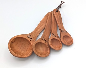 May include: A set of four wooden measuring spoons with a natural light brown wood grain finish. The spoons are connected by a dark brown leather cord. Each spoon is engraved with its measurement: Tbsp, tsp, 1/2 tsp, and 1/4 tsp in black ink.