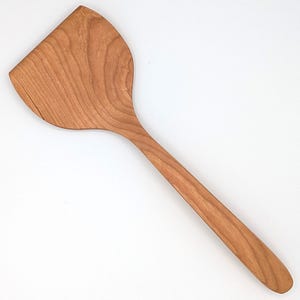 May include: A wooden spatula with a wide, flat, angled head and a long, smooth handle. The utensil features a distinct natural wood grain pattern in shades of light to medium brown, showcasing the organic texture and color variations of the material.