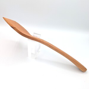 May include: A long-handled wooden cooking spoon with a smooth, light brown grain and a slightly angled, flat-edged bowl. The utensil features a curved handle that tapers toward the end, showcasing the natural texture and warm tones of the wood.