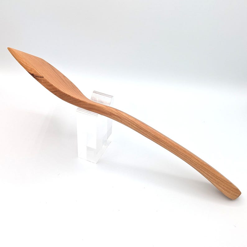 May include: A long-handled wooden cooking spoon with a smooth, light brown grain and a slightly angled, flat-edged bowl. The utensil features a curved handle that tapers toward the end, showcasing the natural texture and warm tones of the wood.