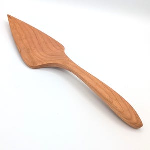May include: A wooden cake server with a triangular blade and a smooth, curved handle. The utensil features a natural light brown wood grain pattern throughout, showcasing a polished finish and a tapered, pointed edge for serving slices.
