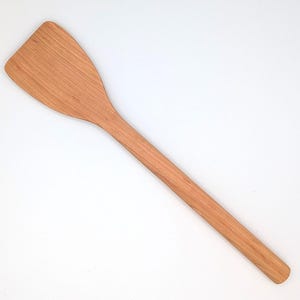 May include: A light brown wooden spatula with a long, straight handle and a wide, flat, angled head. The natural wood grain is visible throughout the surface of the utensil, which features a smooth finish and a rounded end on the handle.