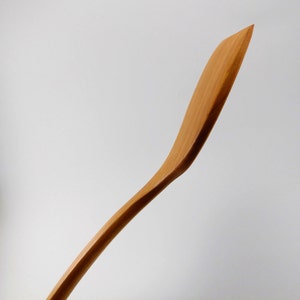 May include: A light brown wooden spatula with a smooth, curved handle and a flat, angled blade. The grain of the wood is visible along the length of the utensil, which features a tapered edge and a slender, ergonomic shape for kitchen use.