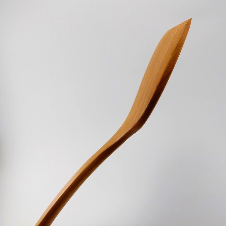 May include: A light brown wooden spatula with a smooth, curved handle and a flat, angled blade. The grain of the wood is visible along the length of the utensil, which features a tapered edge and a slender, ergonomic shape for kitchen use.