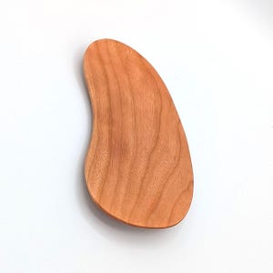 May include: A smooth, light brown wooden tray with a distinct kidney-bean shape. The surface displays a natural wood grain pattern with flowing, organic lines running vertically across the piece, featuring rounded edges and a flat, polished finish.