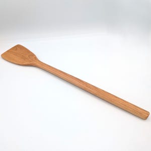 May include: A long-handled wooden spatula with a smooth, light brown grain pattern. The utensil features a flat, slightly angled head that tapers into a rounded, elongated handle, showcasing the natural texture and warm tones of the wood material.