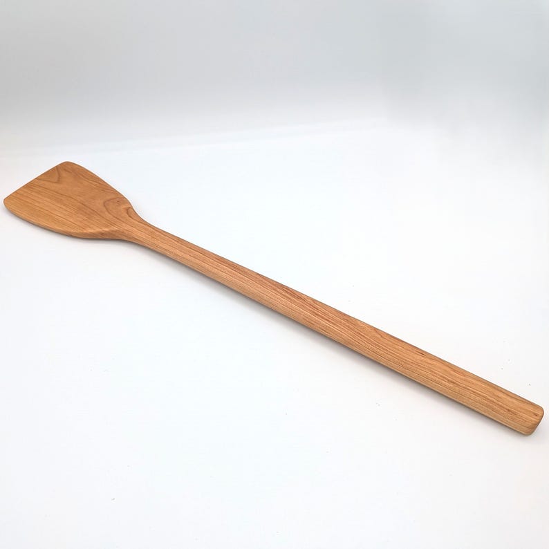 May include: A long-handled wooden spatula with a smooth, light brown grain pattern. The utensil features a flat, slightly angled head that tapers into a rounded, elongated handle, showcasing the natural texture and warm tones of the wood material.