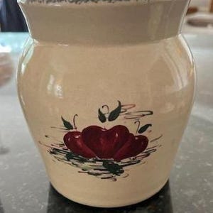 May include: A cream-colored ceramic jar with a wide opening and a slightly flared rim. The jar is decorated with a cluster of three red apples with green leaves and stems. The jar has a glossy finish.
