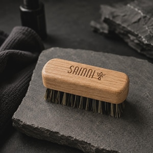 Peut inclure: Une brosse rectangulaire en bois avec des poils foncés et rigides. Le dessus du manche en bois brun clair présente le texte gravé SAINNI suivi d'une petite icône de feuille. La brosse a un design aux bords arrondis et une finition en bois naturel.