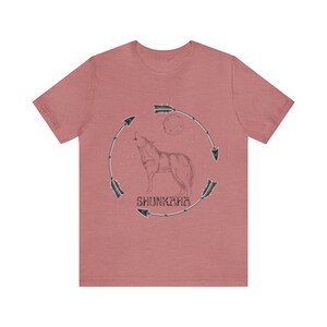 Puede incluir: Una camiseta de color rojo heather con una ilustraci&oacute;n en blanco y negro de un lobo aullando a la luna. El lobo est&aacute; rodeado por un c&iacute;rculo de flechas y la palabra "SHUNKARA" est&aacute; escrita debajo del lobo.