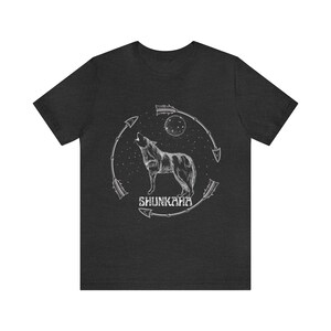 Puede incluir: Una camiseta gris oscuro jaspeada con un gr&aacute;fico blanco de un lobo aullando a la luna. El lobo est&aacute; rodeado de flechas y el texto "SHUNKAHA" est&aacute; debajo del lobo.