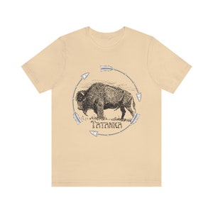 Pu&ograve; includere: Una t-shirt beige con un'illustrazione in bianco e nero di un bufalo circondato da frecce. Il testo "Tatanka" &egrave; stampato sotto il bufalo.